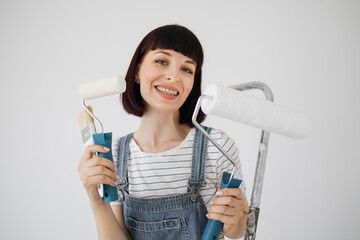 Beautiful young woman at age of 30, sitting on ladder and holding paint rollers and brush in hands with which she has just painted wall in living room.