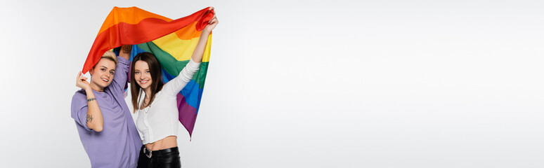 Fototapeta premium young and joyful lesbians with lgbtq flag looking at camera isolated on grey, banner.