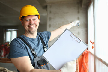 Smiling builder worker in hard hat holding blank sheet of paper on clipboard