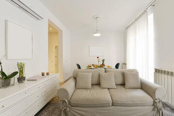 Room with a two-seater sofa with gray fabric, windows with white curtains and white wooden furniture