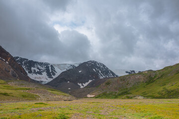 Dramatic alpine scenery with green mountain valley and snow mountains under cloudy sky in changeable weather. Atmospheric overcast landscape with snow mountain range and green valley in sunlight.