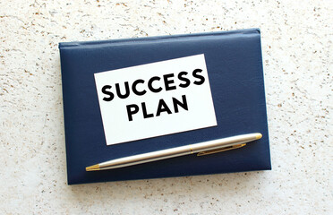 Text SUCCESS PLAN on a business card lying on a blue notebook next to the pen.