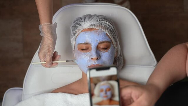 Female beauty business owner making promo photos of woman's client face with cosmetic skincare product on by smartphone for social medias and promotions. Beauty industry, spa and skincare concept.