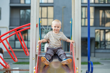 Fototapeta na wymiar Cute two year old boy plays on playground slide on summer. Active kid on colorful slide and swing.