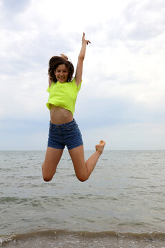 Young slender teenage girl jumps for happiness on the seashore in summer with jeans shorts
