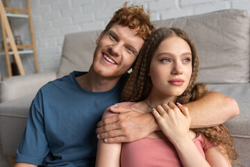 redhead and happy young man smiling and hugging pretty girlfriend in living room.