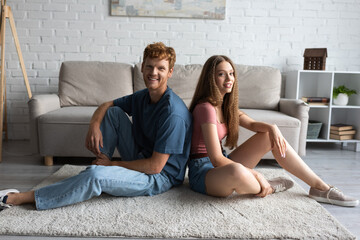 full length of happy young couple sitting on carpet and looking at camera in living room.