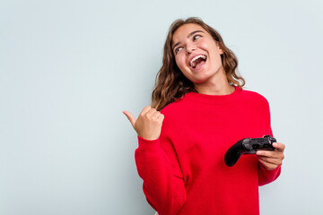 Young caucasian gamer woman holding a game controller isolated on blue background points with thumb...