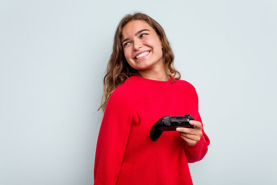 Young Caucasian Gamer Woman Holding A Game Controller Isolated On Blue Background Looks Aside Smiling, Cheerful And Pleasant.