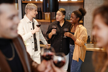 Group of young intercultural friends with glasses of red wine chatting to one another in the kitchen while enjoying home party
