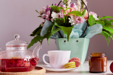 Autumn tea time - cup of tea with macarons, glass teapot, candle. Herbal raspberry natural tea. Cozy home with bouquet of flowers, tea and dessert. Self care, wellness lifestyle.