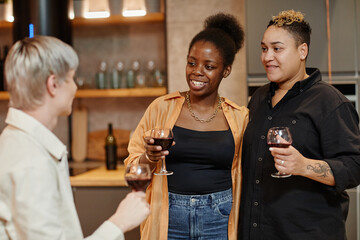 Happy young lesbian couple with glasses of red wine chatting to their friend while standing in front of him in the kitchen at home party