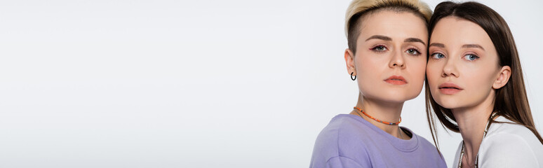 blonde woman with makeup looking at camera near young brunette girlfriend isolated on grey, banner.