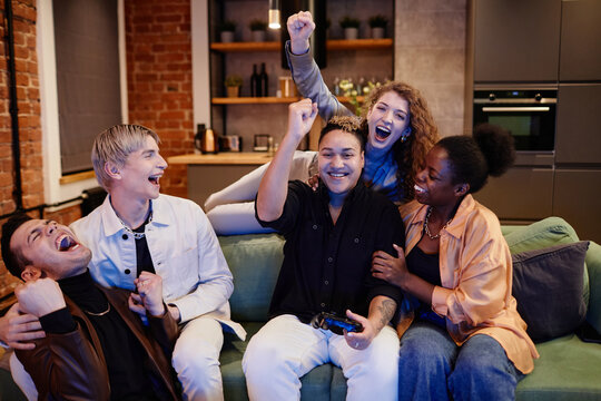 Intercultural ecstatic friends expressing triumph while relaxing on couch in living room together on weekend and playing video game