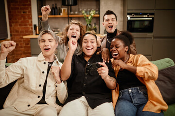 Group of young intercultural ecstatic friends shouting while sitting on couch in living room on weekend and playing video game