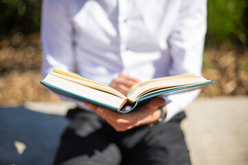 Closeup view of young man reading the bible. Young man with the bible at a park sitting on a bench. Horizontal.