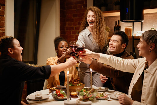 Young ecstatic friends clinking with red wine in wineglasses over dinner table served with homemade food and decorated with burning candles - Powered by Adobe