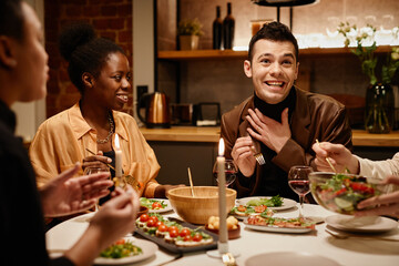 Happy young grateful man saying thanks to woman who prepared tasty food while sitting by served dinner table among friends