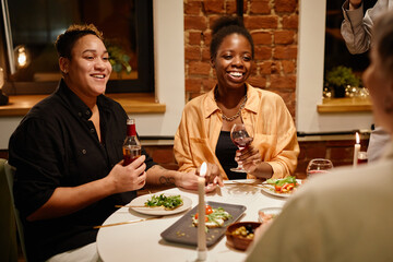 Happy young lesbian couple with alcoholic drinks sitting by served table and talking to friends who came to dinner and home party