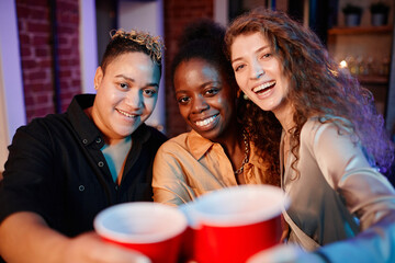 Cheerful pretty girls clinking with plastic glasses and looking at camera while standing in home environment and enjoying party