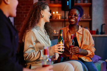 Three cheerful intercultural girls with bottles of beer having rest on couch and chatting while enjoying home party on weekend evening