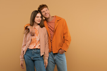 Fototapeta premium redhead young man standing with hand in pocket and hugging cheerful girlfriend isolated on beige.
