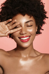 Vertical shot of smiling African American girl, has glowing perfect facial skin, body after shower, using cleansing gel, nourishing cream cosmetic, pink background