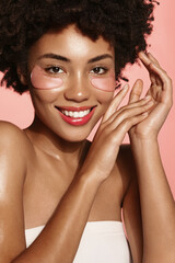 Vertical shot of beauty woman, african american girl puts on hyaluron under eye patches from dark circles, stands over pink background