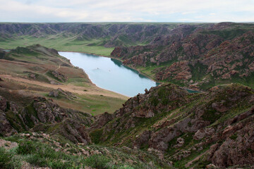 Fototapeta premium Top view of a wide river in a mountain valley, cloudy summer day, the sky is in the clouds, the mountain slopes are covered with rocks and green grass, there is sand on the river bank, Ili river, Kaza