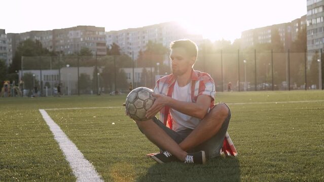 Frustrated man sits on football field squeezes punctured ball.
