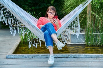 Middle aged woman with smartphone sitting in hammock