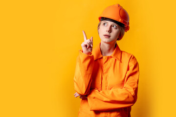 Surprised blond hair worker woman in orange helmet and suit on yellow background