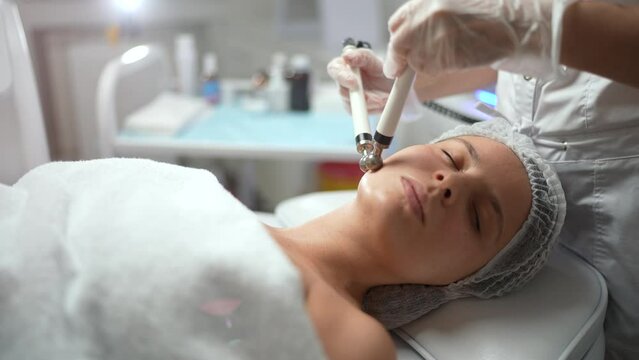 Beautiful young female receiving facial microcurrent therapy at spa salon. Professional skincare, cosmetology, beauty treatment concept. High quality 4K footage