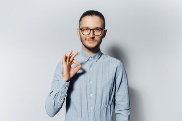 Studio portrait of young confident guy showing ok gesture on white background. Wearing blue shirt and eyeglasses.
