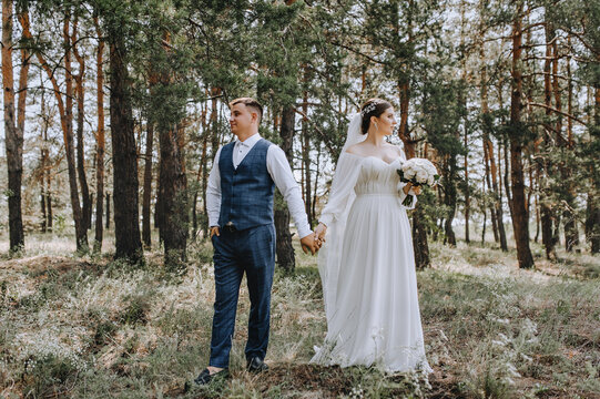 Beautiful newlyweds stand in the forest in nature, holding hands. Close-up wedding portrait, photo of stylish groom and young brunette bride in white long dress with bouquet of roses.