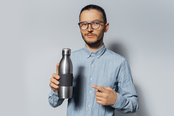 Studio portrait of young man pointing finger on metal bottle in his hand, on white background, wearing eyeglasses and blue shirt.