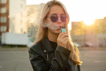 Young woman with sunglasses smoke vape pod system on a city street. Female inhales and exhales vapor of electronic cigarette.