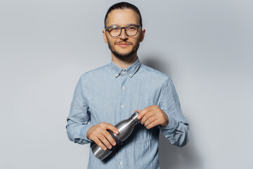 Studio portrait of young man holding metal bottle in hand on white background, wearing eyeglasses and blue shirt.