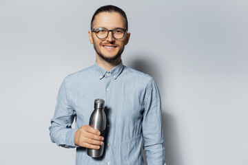 Studio portrait of young smiling guy holding metal bottle in hand on white background, wearing eyeglasses and blue shirt.