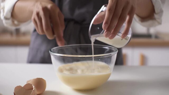 Female hands add milk to bowl of beaten eggs with mixer on wooden kitchen table. Waffle cookie recipe step by step. Hand mixer beats eggs, milk. Beating dough with mixer. Whipping dough for pancakes
