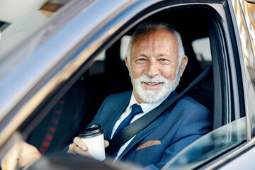 An old businessman drives a car. A happy senior businessman sits in his car, driving himself to work and holding takeaway coffee while looking through the window.