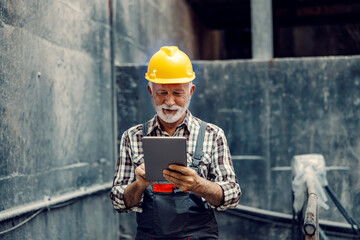Factory management and heavy industry operating. A smiling senior factory worker with a helmet on his head stands in a metal construction facility and uses a tablet to make the job easier.