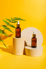 Glass Bottles of serum oil cosmetic product, branch fern on podium pedestal on yellow background. Natural skin care beauty product concept. Mockup for branding