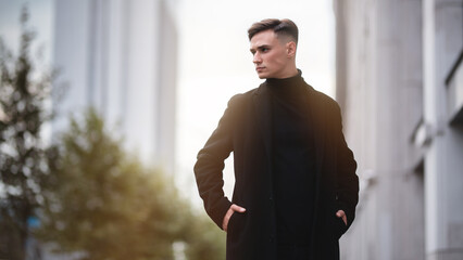 Young attractive man in stylish black clothes on the street among the buildings. Business concept. The guy walks down the street against the backdrop of skyscrapers and office buildings © pridneprovskiy