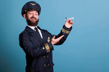 Smiling pilot with uniform playing with airplane model side view, aviation academy aviator holding commercial plane toy. Funny aircraft capitan, studio medium shot on blue background © DC Studio