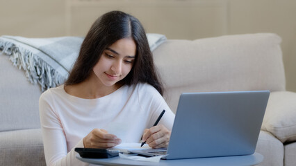 Young housewife arab middle eastern woman considers utilities uses calculator control domestic budget counts incomes outcomes bills writes notes documentation make paperwork account for spending costs
