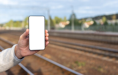 Hand holding phone mockup on railway background with copy space. Mobile application with online train timetable. Smartphone template with empty display