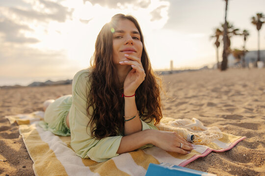 Image of happy smiling fair-sjinned woman keep hand on chin look to camera. Lying at beach in morning with book. Rest time concept 