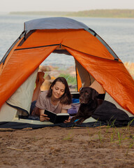 woman and a Labrador retriever dog on a camping trip in nature. a woman reads a book lying in a...