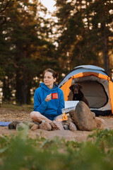 woman and a Labrador retriever dog on a camping trip with a tent in nature. a woman is reading a book or drinking tea from a red cup next to a campfire. a pet on vacation with his family, traveling